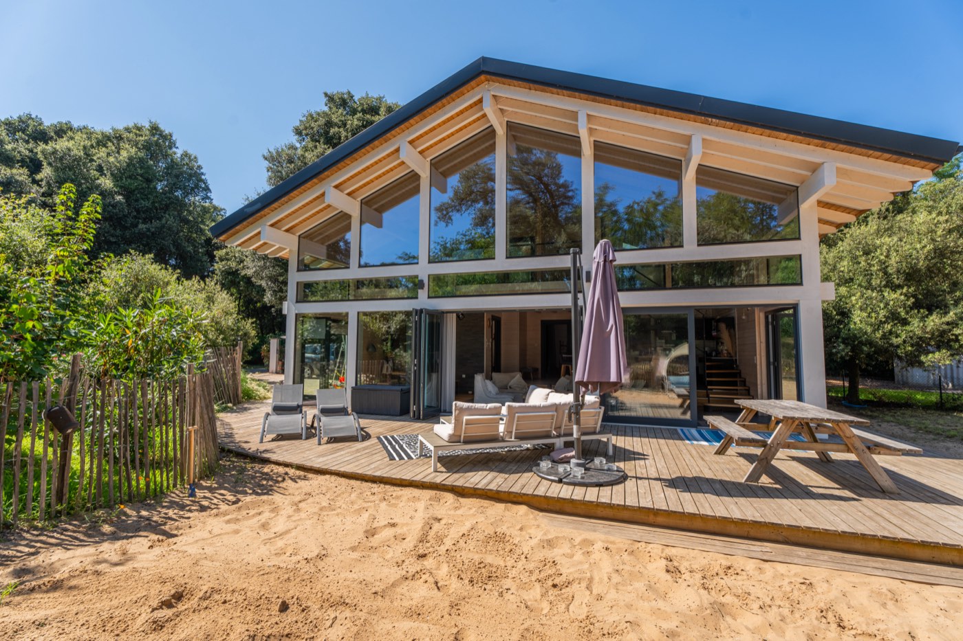 Le Chalet Moana vu de la terrasse : façade vitrée toute hauteur, bains de soleil, parasol et table de pique-nique, le sable au pied du plancher